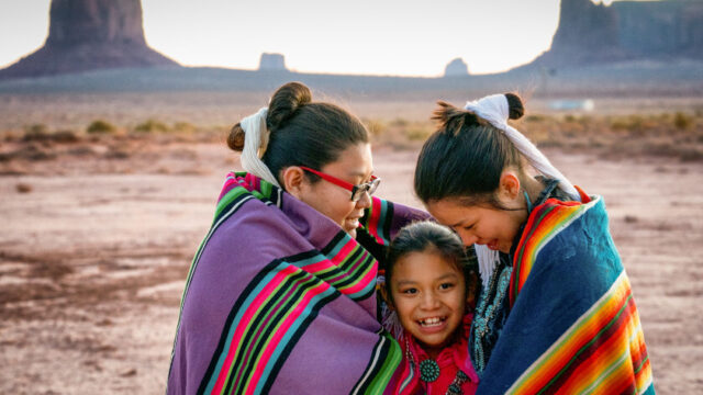 In Monument Valley, two older Navajo sisters face inward and look at their younger sister, who is in between them. All three are smiling and in traditional dress, with handwoven traditional blankets around their shoulders.
