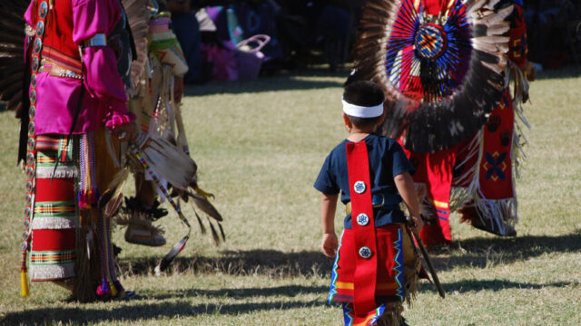 A small child in traditional Native American attire walks among adults who are also dressed in traditional Native American attire