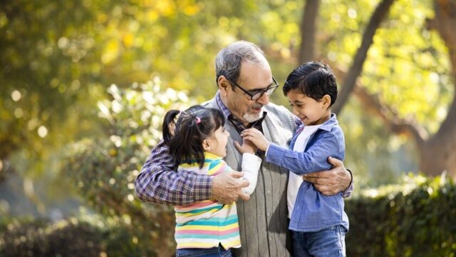 In a park, an Indian grandfather kneels to embrace his young granddaughter and grandson, who are both smiling and placing their hands on his heart