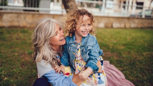 A white grandmother and her young granddaughter smile as the grandmother sits on the ground and hugs her standing granddaughter