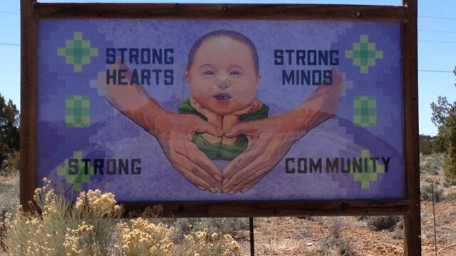 Photograph of a billboard with a painting of a Native child behind adult hands making a heart shape. The billboard says, 