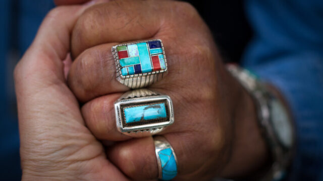 A Native American elder's hand, with traditional turquoise rings, clasps the hand of another person
