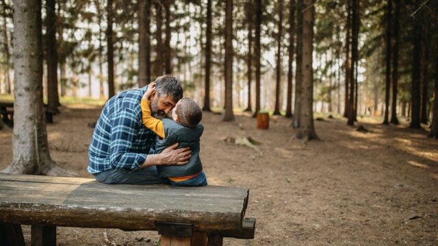A older man and a little boy sit on a picnic table in the woods with their arms around each other, with the man bending and the little boy reaching up