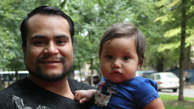 A Native man holds a baby/toddler in his arm and both look at the camera