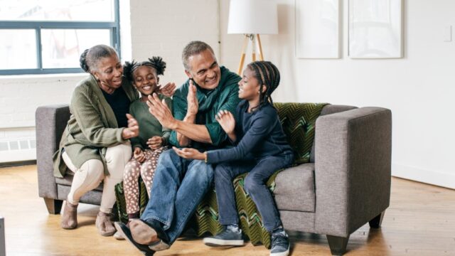 A Black grandfamily sits on a couch and members of the family look at each other and give each other high-fives. Depicted are a grandmother, a grandfather, an elementary-age girl, and an elementary-age boy.