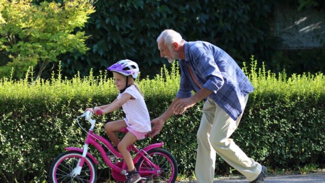 A little girl learns to ride a bike. Depicted are the girl, who is wearing a helmet and riding her bike, and her grandfather, who is holding the back of her seat. Both individuals are white.