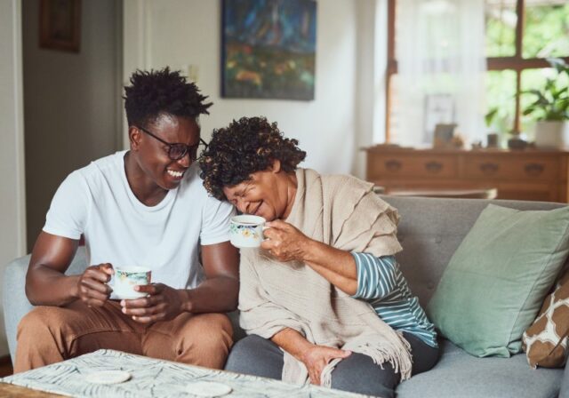 A grandmother and her teenage grandson sit on the couch with coffee mugs. The grandson smiles at the grandmother, who is laughing with her head on his shoulder. Both individuals are Black.