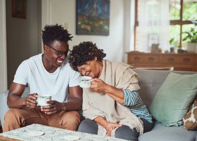 A grandmother and her teenage grandson sit on the couch with coffee mugs. The grandson smiles at the grandmother, who is laughing with her head on his shoulder. Both individuals are Black.