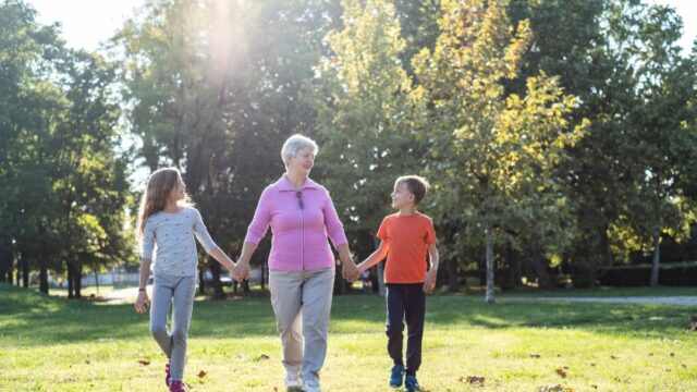 A white grandmother holds hands with her granddaughter and grandson and walks in a field, with trees behind them. All three are looking at each other.