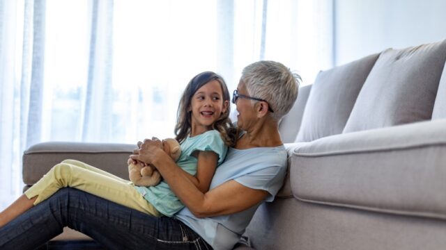A white grandmother sits on a pillow on the floor and leans against a couch, with her legs stretched out in front of her and her arms around her elementary-aged granddaughter, who is in her lap. The two individuals are looking at each other, and the girl is holding a teddy bear.