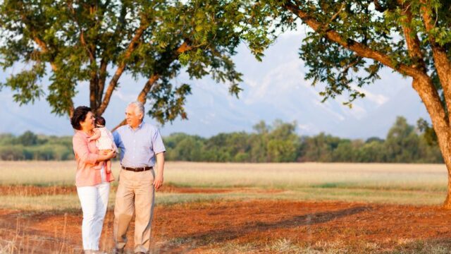 A grandmother and grandfather walk together and look at each other. The grandmother holds their baby granddaughter, and the grandfather has his arm around the grandmother. The family is Latinx.