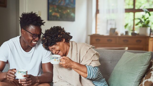 A grandmother and her teenage grandson sit on the couch with coffee mugs. The grandson smiles at the grandmother, who is laughing with her head on his shoulder. Both individuals are Black.