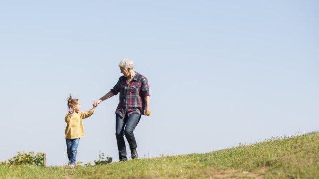 A young girl and her grandmother hold hands and walk down a hill in a grassy field. Both individuals are white.