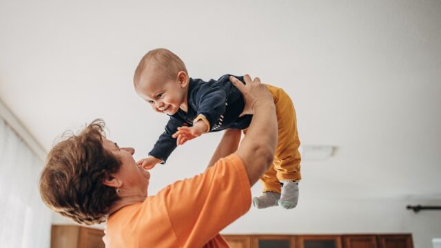 A smiling white grandmother looks at her baby grandson as she holds him in the air above her head. The grandson smiles and reaches his hands out towards his grandmother.