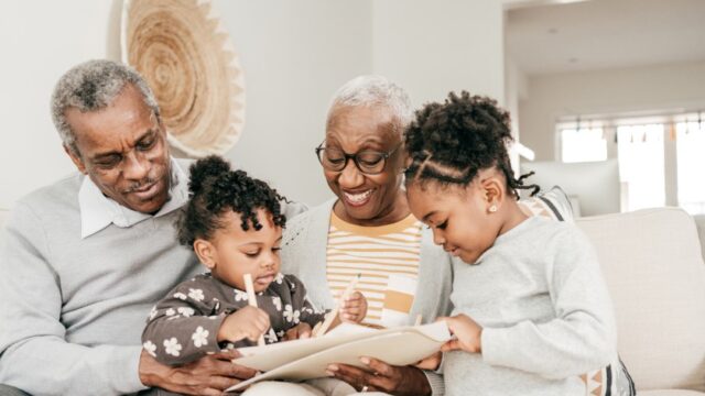 A Black grandmother and grandfather sit with their two young granddaughters on a couch and watch as the granddaughters look intently at papers and color/write on them. The grandmother is smiling..
