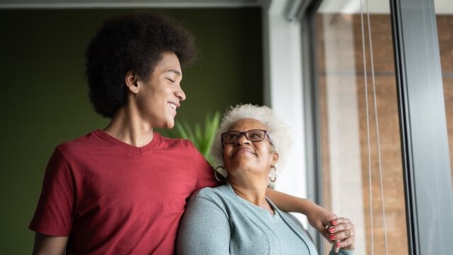 A Black teenage boy stands with his arm around his grandmother and looks at her. She holds his hand that is draped over her shoulder and looks back at him.