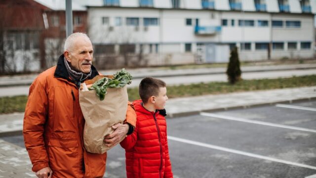A white grandfather holds a cane in one hand and a bag of groceries in the other as he crosses a parking lot with his grandson.
