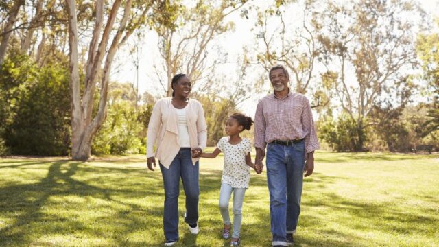 A grandmother, an elementary school-aged girl, and a grandfather hold hands and walk together in a park. The grandmother and grandfather are smiling, and the girl is looking off to the side. All three individuals are Black.