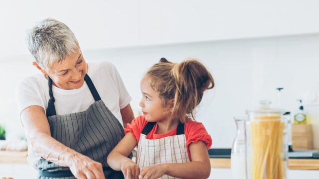 A grandmother and granddaughter look at each other as they cook/bake together.