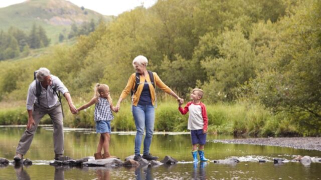 A white grandfather, granddaughter, grandmother, and grandson look at each other and hold hands as they balance on rocks in water.