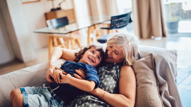 A white grandmother and her young grandson smile and look at each other as the grandmother holds and hugs her grandson as they recline on the couch together.