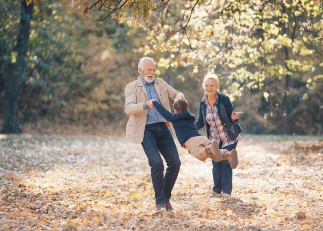 A white grandfather swings his young grandson by the arms in a park on a fall day. The grandmother smiles and leans in towards her husband and grandson.