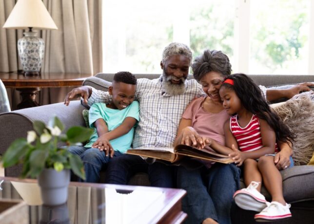 A Black family, consisting of a grandson, and grandfather, a grandmother, and a granddaughter, sit together on a couch and smile as they look through a book or photo album together.