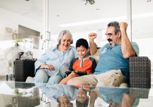 A Hispanic/Latino grandmother, grandson, and grandfather play a video game together on the couch, and the grandfather pumps his arms in celebration.