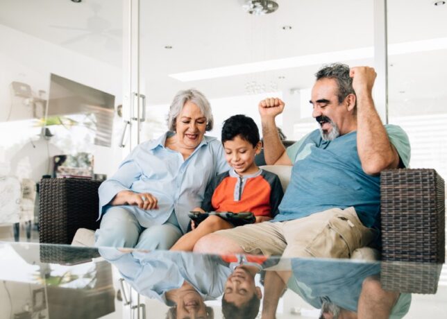 A Hispanic/Latino grandmother, grandson, and grandfather play a video game together on the couch, and the grandfather pumps his arms in celebration.