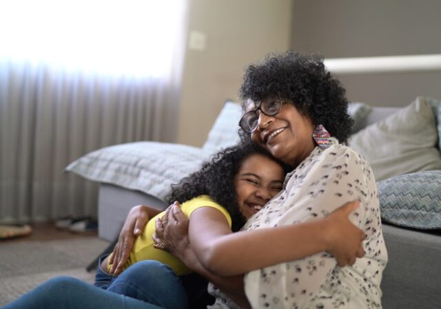 A Black grandmother and granddaughter sit and smile as they hug each other.