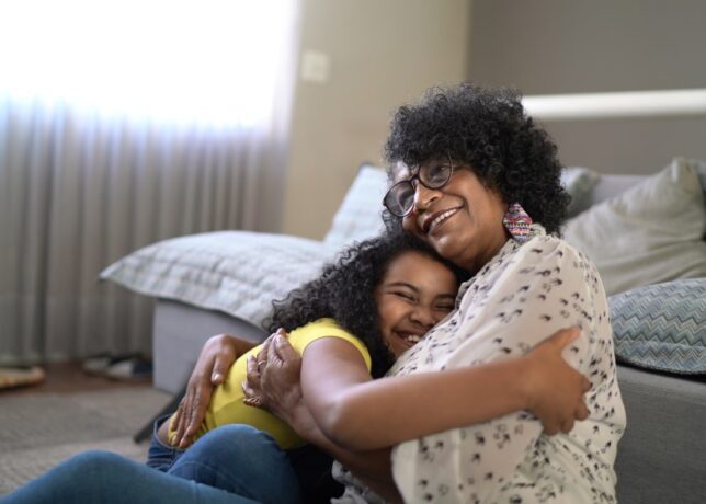 A Black grandmother and granddaughter sit and smile as they hug each other.