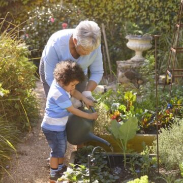 A grandmother and her young grandson water the garden together. Both the grandmother and the grandson are Black.