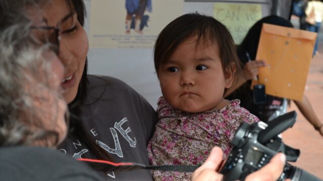 A Native woman holds a baby as an older Native woman shows her a photo on a digital camera