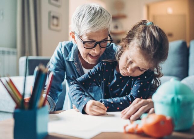 A white grandmother smiles and leans over her young granddaughter's shoulder as the child writes/colors