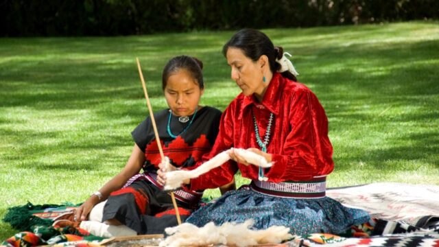 A Navajo woman and child sit outside on a blanket and the woman shows the child how to weave