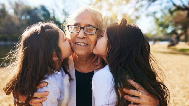 A Hispanic/Latina grandmother smiles as two granddaughters each kiss her on the cheek. All three are outside.