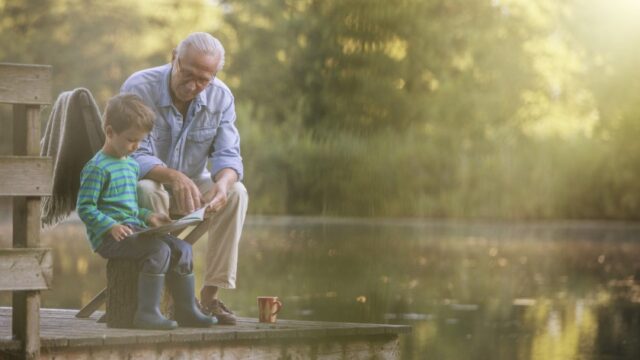 A white grandfather and his young grandson sit on a wooden dock, surrounded by water and trees