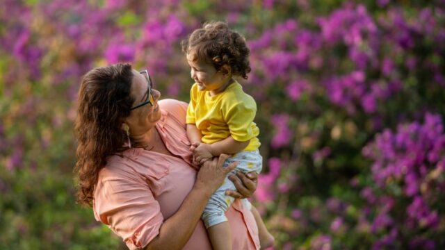 A Hispanic/Latina woman holds her young grandchild outside and the two smile at each other. Behind them is a bush in bloom.