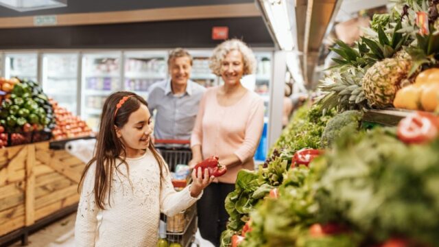 A young white girl picks out a red pepper in the grocery store as her grandparents smile and watch.