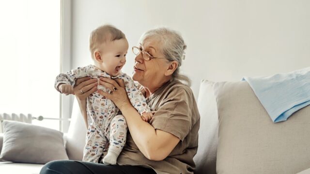 A white grandmother sits on a couch and holds her baby grandchild in a standing position on her lap. The grandmother is looking at the baby and the baby is smiling.