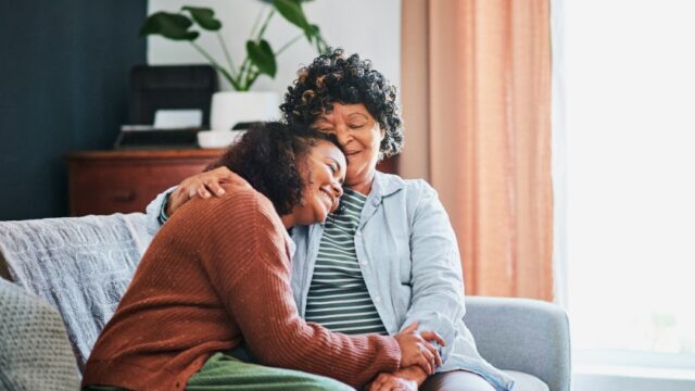 A young woman and her grandmother smile as they sit on the couch together, and the young woman leans her head on her grandmother’s shoulder. Both individuals are Black.