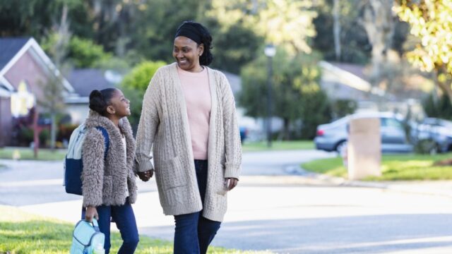 A Black grandmother and granddaughter look at each other and smile as they walk to school together. They are holding hands.