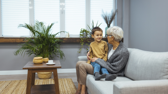 A white grandmother sits on a couch with your young grandson sitting sideways on her lap, with his legs stretched out over her legs. Her arms are around him and hey are looking at each other.