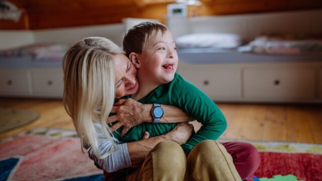 A white grandmother sits on the floor with her grandson, who has Down Syndrome, and hugs him closely while he smiles