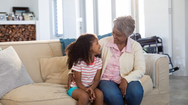A Black grandmother and granddaughter sit on a couch together and smile at each other. There is a wheelchair in the background of the image.