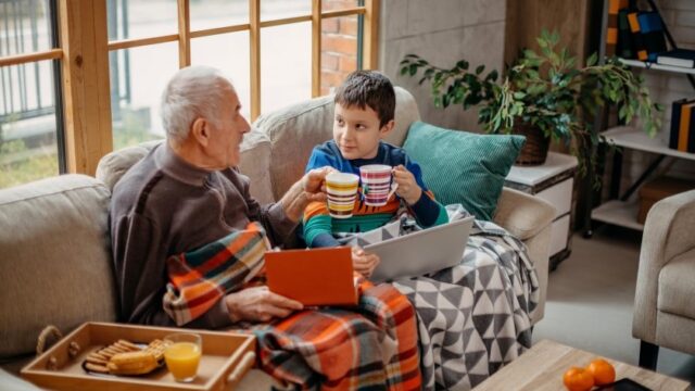 A white grandfather and grandson sit on the couch together, with blankets over their legs, and look at each other as they clink mugs