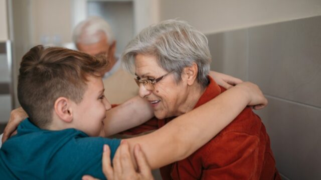 A white grandson stands with his arms on his grandmother's shoulders. She has her hand on his arm, and they are smiling at each other. The grandfather is in the background.