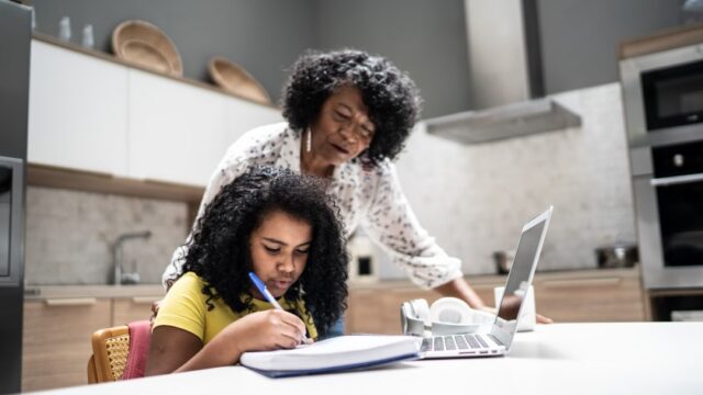 A Black grandmother looks over her granddaughter's shoulder as her granddaughter works on homework.