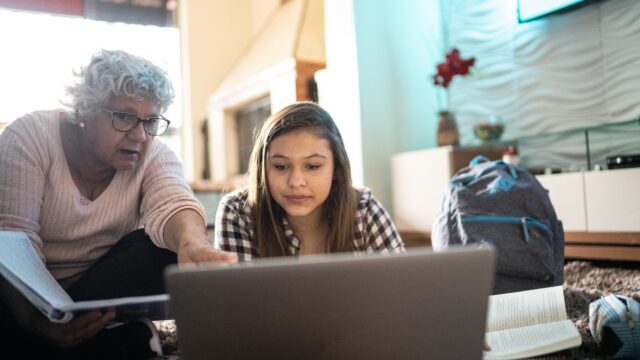 A Latina grandmother helps her teenage granddaughter with homework, pointing to something on a laptop screen