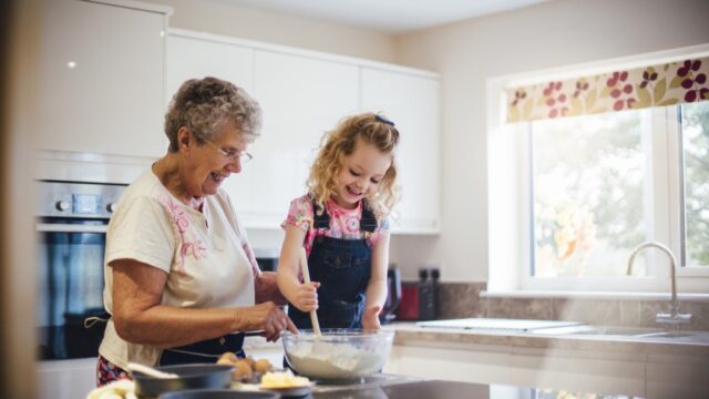 A white grandmother and her young granddaughter smile as they mix ingredients together in the kitchen.